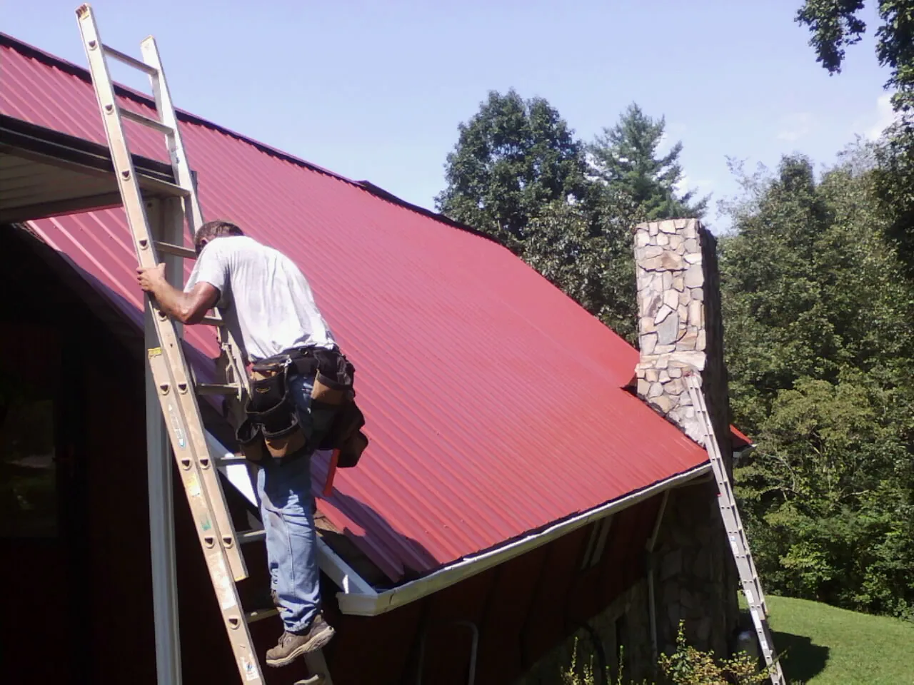 Roofer on a ladder beside a red metal roof and stone chimney — Soffit & Fascia Repair by Roofing Brothers in Dornsife